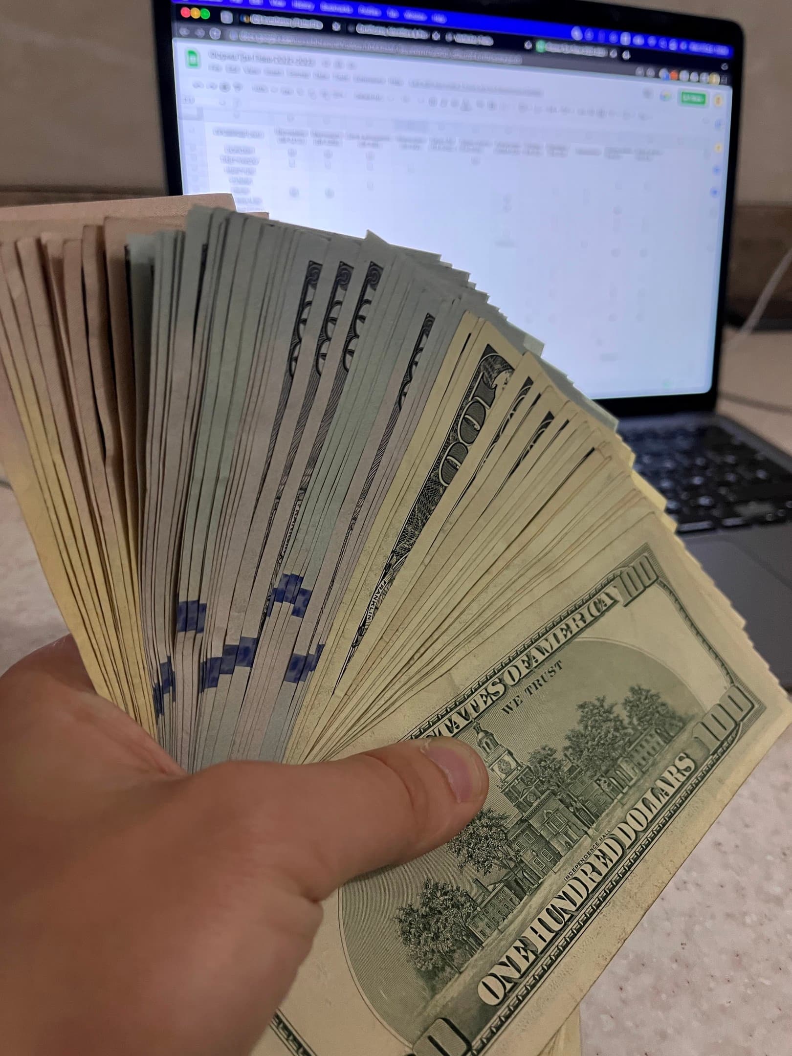 A fan of hundred-dollar bills in front of a laptop spreadsheet.